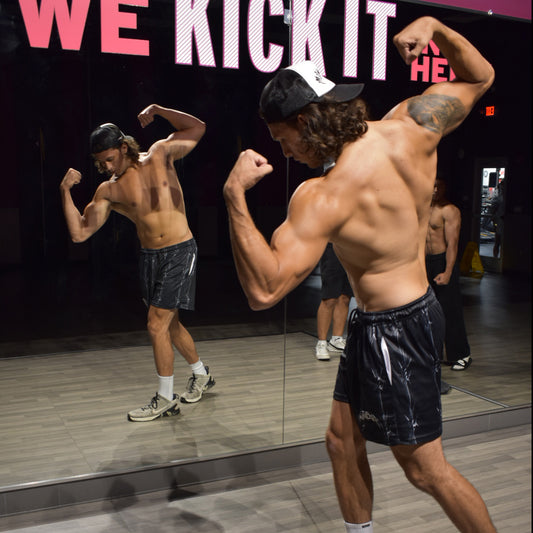 Joshua flexing muscles in front of a mirror with 'WE KICK IT' text on a black background 